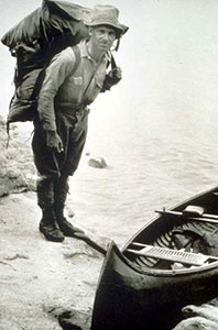 Arthur Carhart carrying a heavy backpack next to a canoe